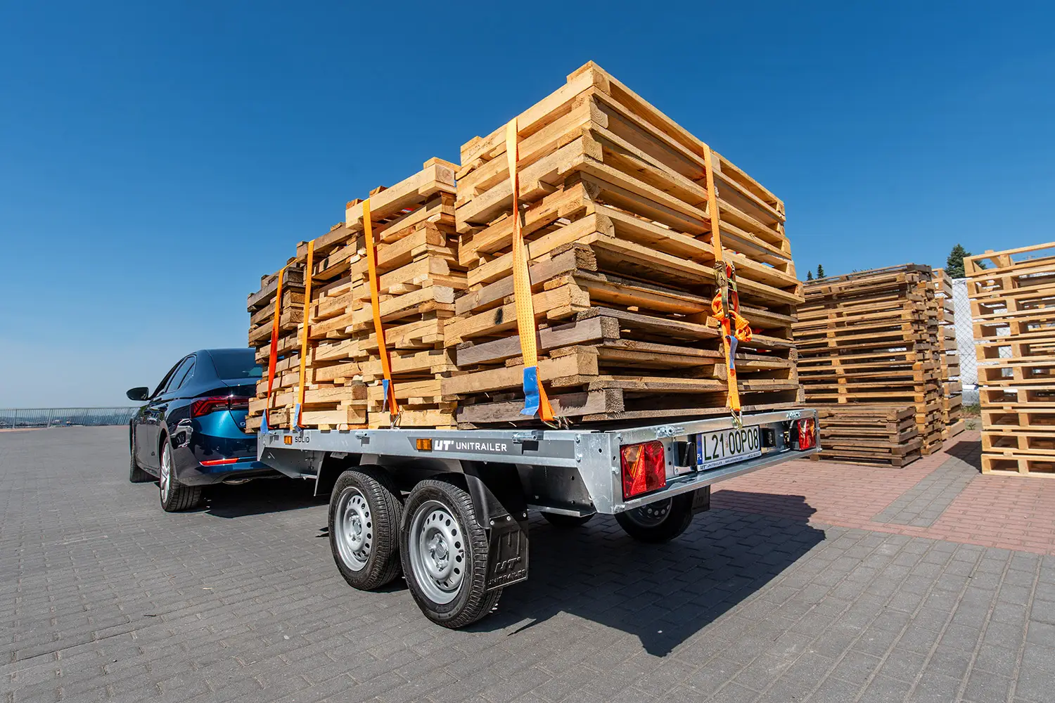 Remorque double essieu Unitrailer attelée à une voiture sombre, chargée d'une haute pile de palettes en bois sécurisées par des sangles de transport.