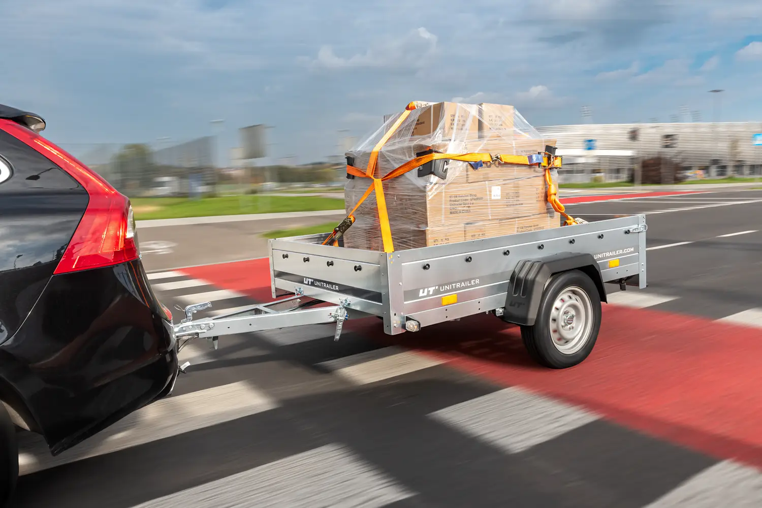 Remorque Unitrailer attachée à une voiture noire, transportant des cartons emballés sous film étirable et sécurisés par des sangles d'arrimage pendant la conduite.