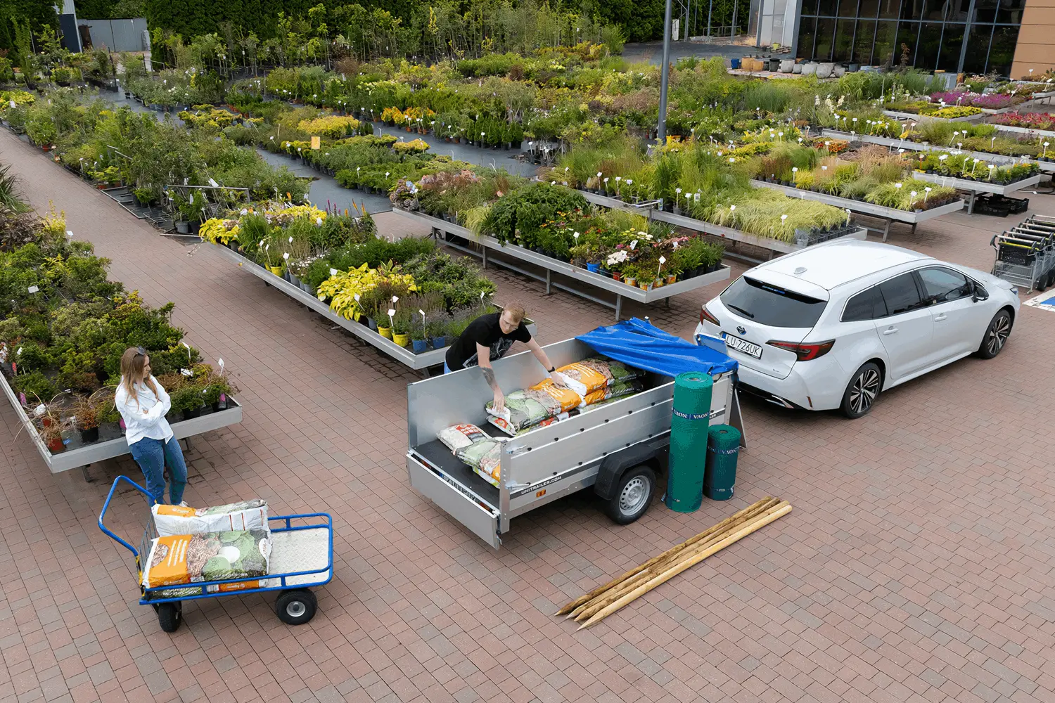 Vue d'en haut d'une jardinerie où une voiture blanche avec une remorque Unitrailer est chargée de sacs de terreau et de fournitures de jardinage.